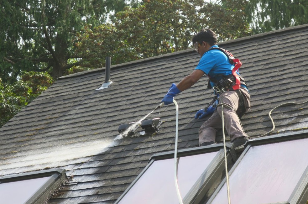 Technician pressure-washing a shingled roof above skylights with safety harness